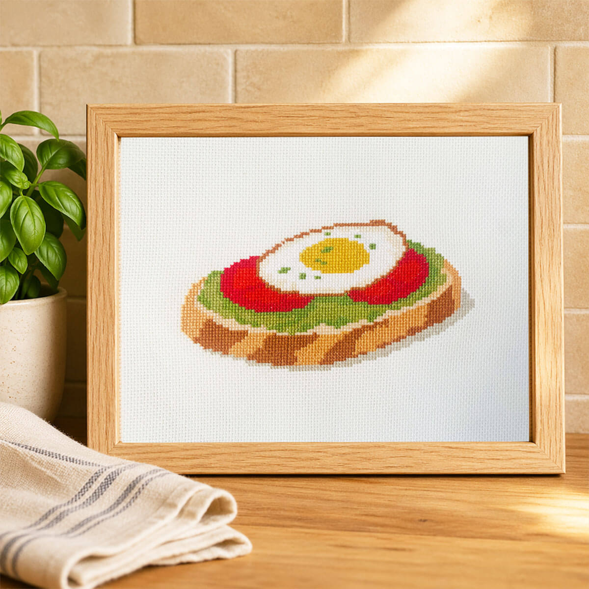 Framed cross-stitched picture of an avocado toast with an egg on a wooden surface.