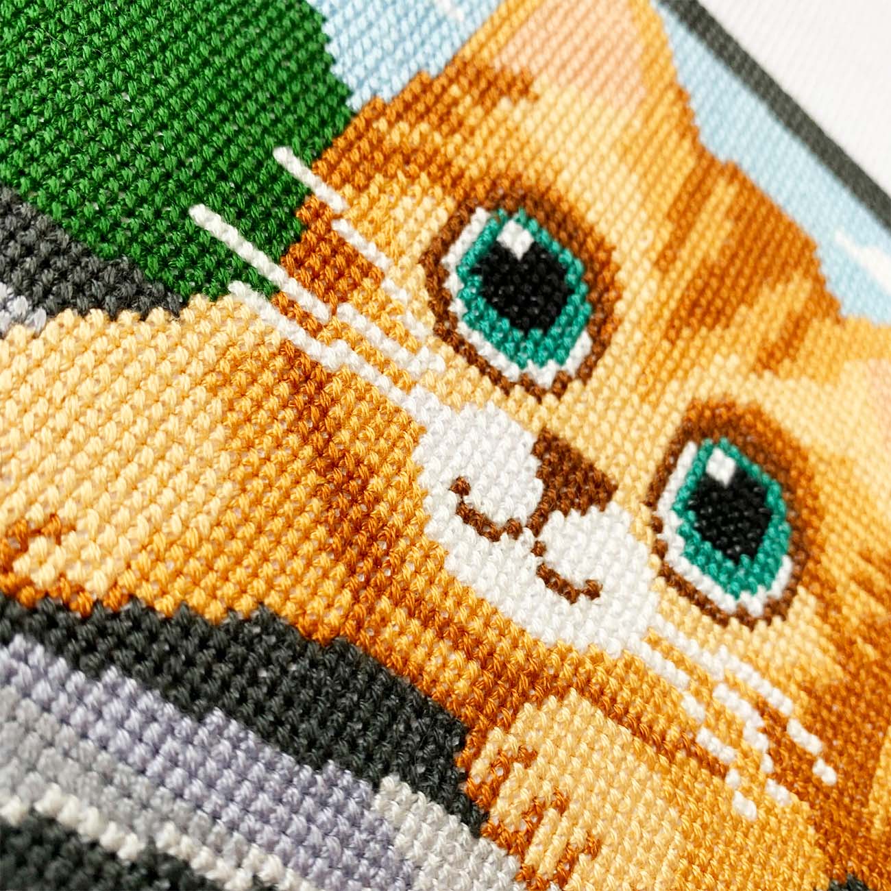 Close-up of a cross-stitched orange cat with green eyes on fabric.