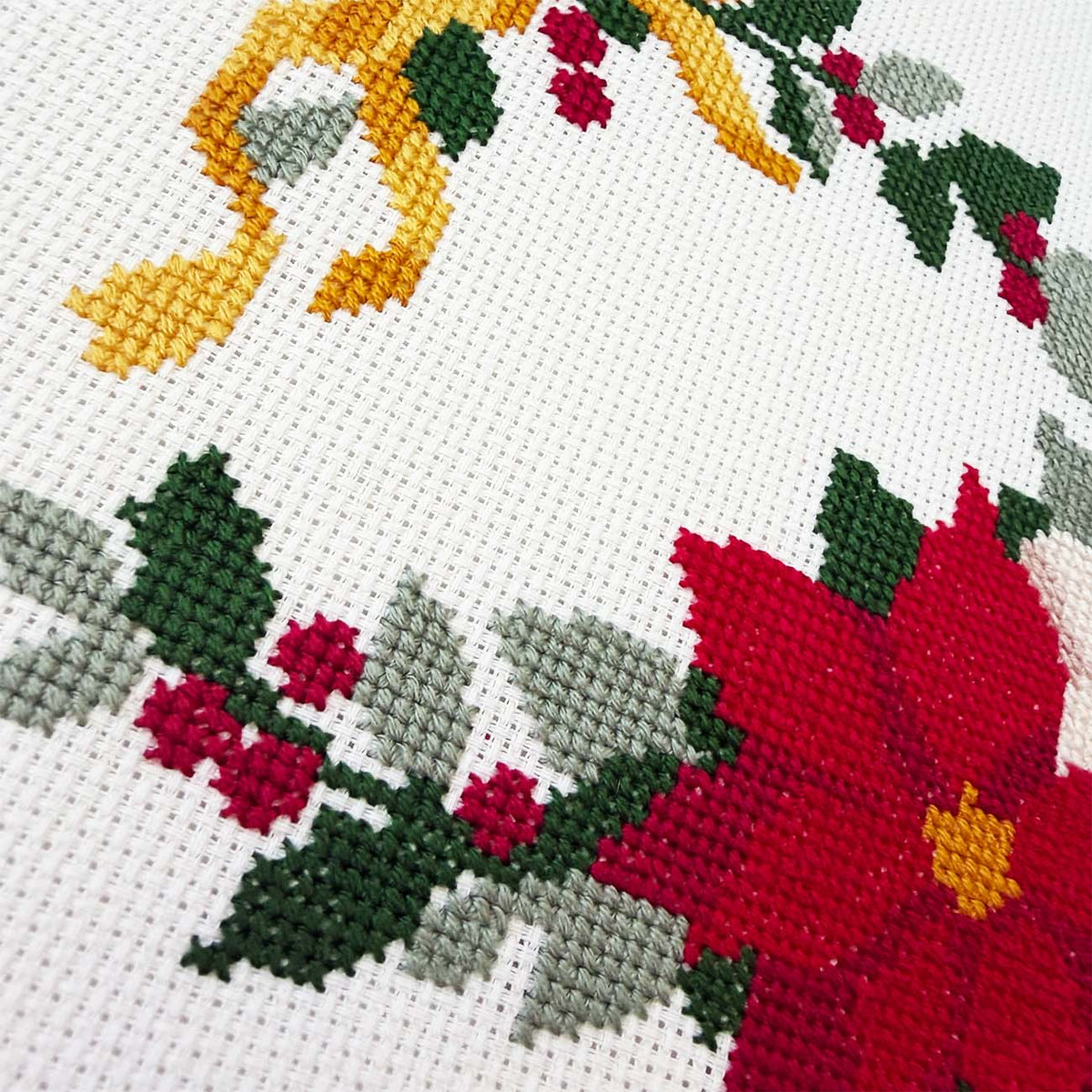 Close-up of a cross-stitch pattern on off-white Aida fabric, featuring a large red flower with yellow center, green leaves, and small red berries.