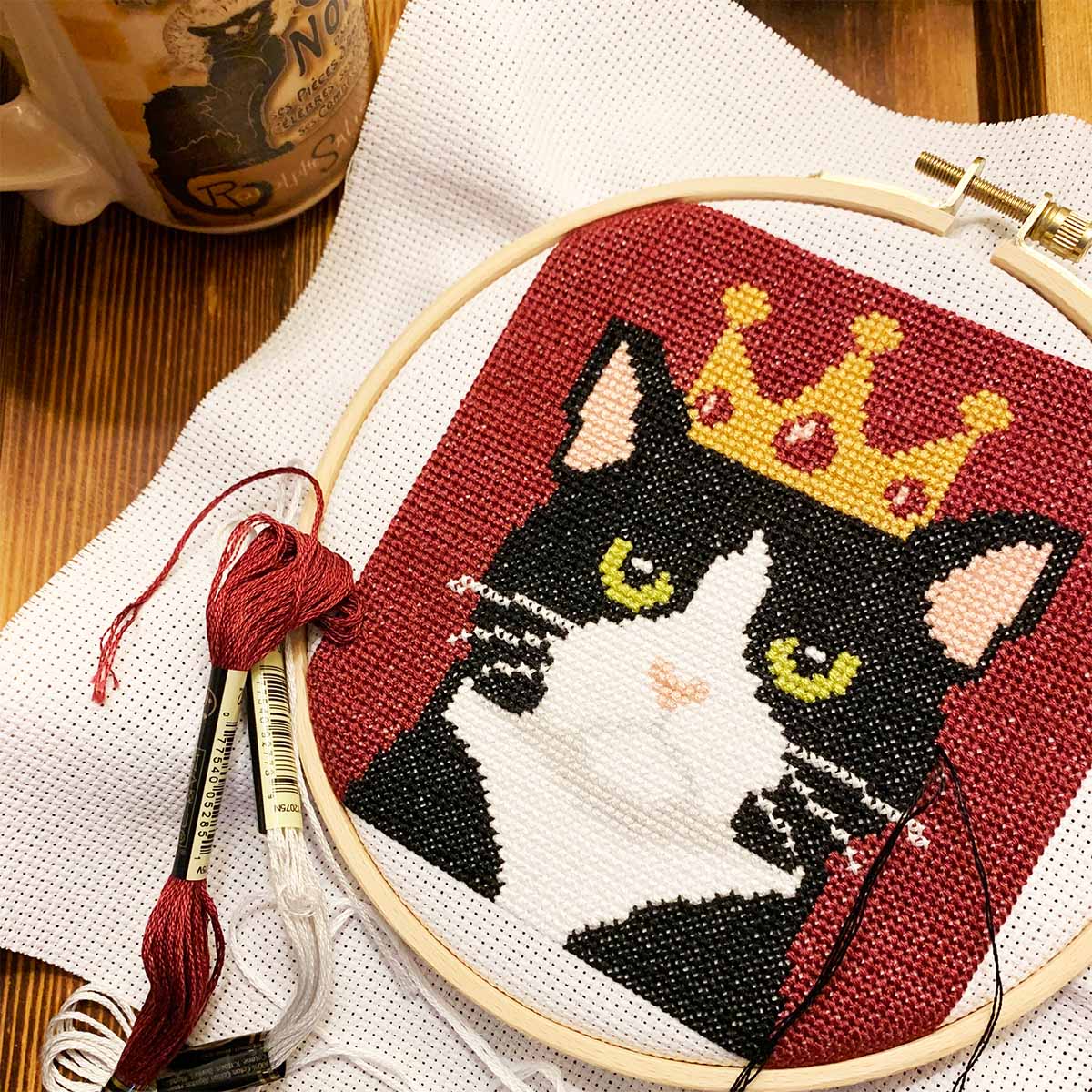 Embroidery hoop with a black and white cat design wearing a crown, on a wooden surface.