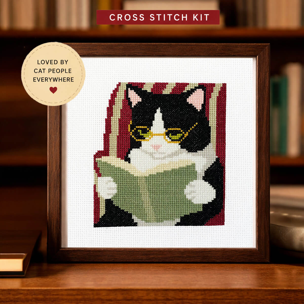 Cross stitch kit with a cat reading a book design, framed on a wooden surface.