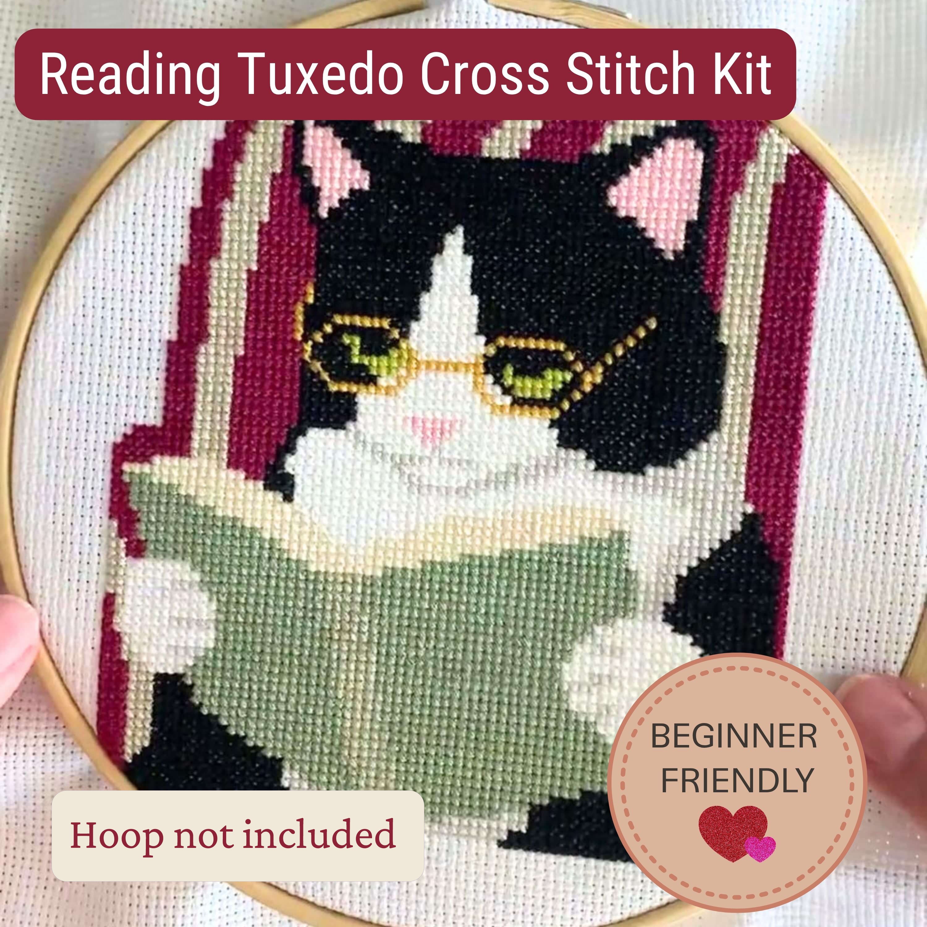 A completed cross stitch design of a black-and-white cat reading a green book, shown inside a hoop. Text banners read 'Reading Tuxedo Cross Stitch Kit' and 'Beginner Friendly,' with one banner noting 'Hoop not included'.