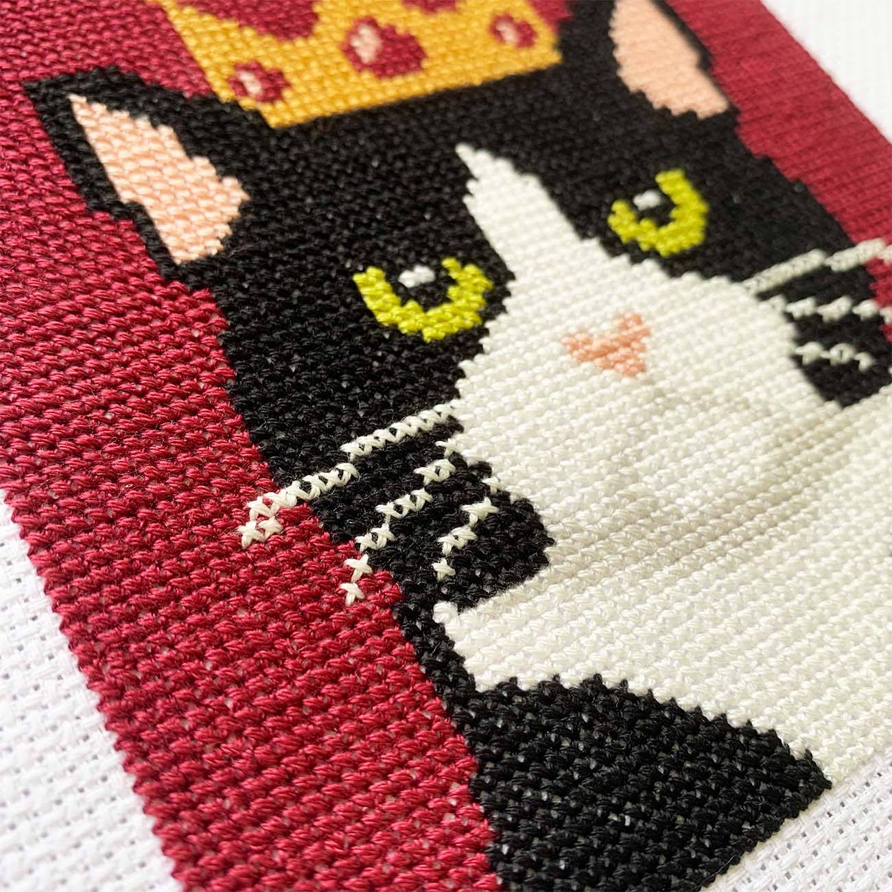 Close-up stitches of a cross-stitched design of a black and white cat with a yellow crown on a red background showing thread texture and color details.