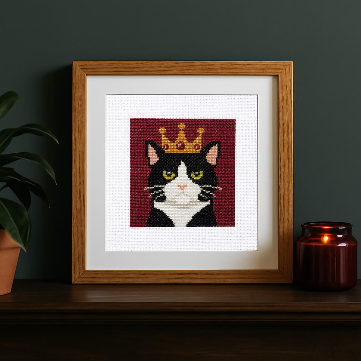 Framed cross-stitched picture of a cat with a crown on a dark surface with a plant and candle.