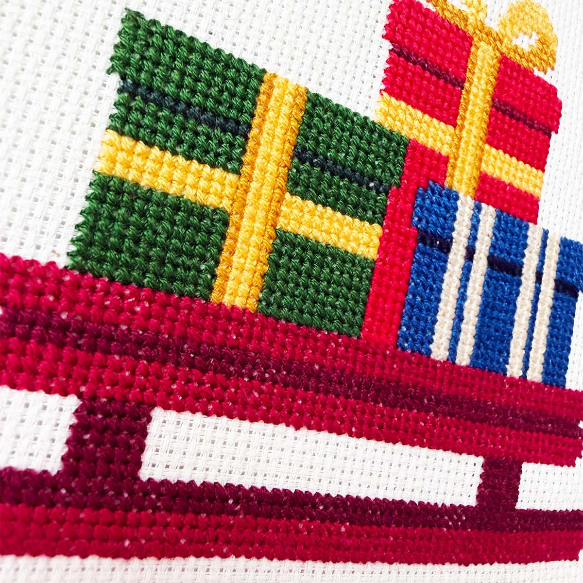 Close-up of a cross-stitch design depicting colorful Christmas presents on a red sleigh over an off-white canvas background.