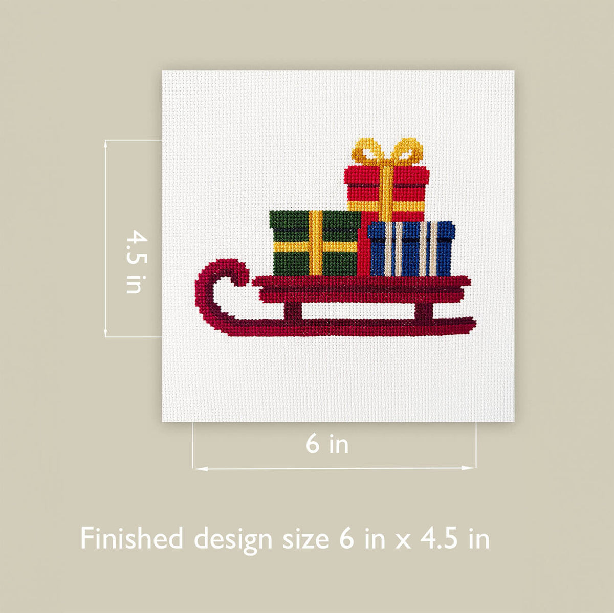 A cross-stitch design of a red sleigh carrying colorful wrapped gifts, shown on a white fabric square with dimension markers indicating 6 in by 4.5 in, on a beige background with text reading 'Finished design size 6 in x 4.