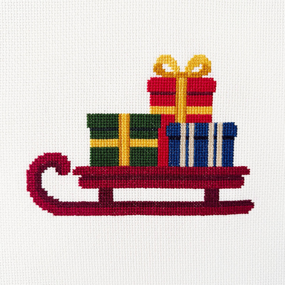 A festive cross-stitch design featuring a red sled carrying three wrapped Christmas presents in green, red, and blue, with yellow ribbons and bows, on an off-white Aida background.
