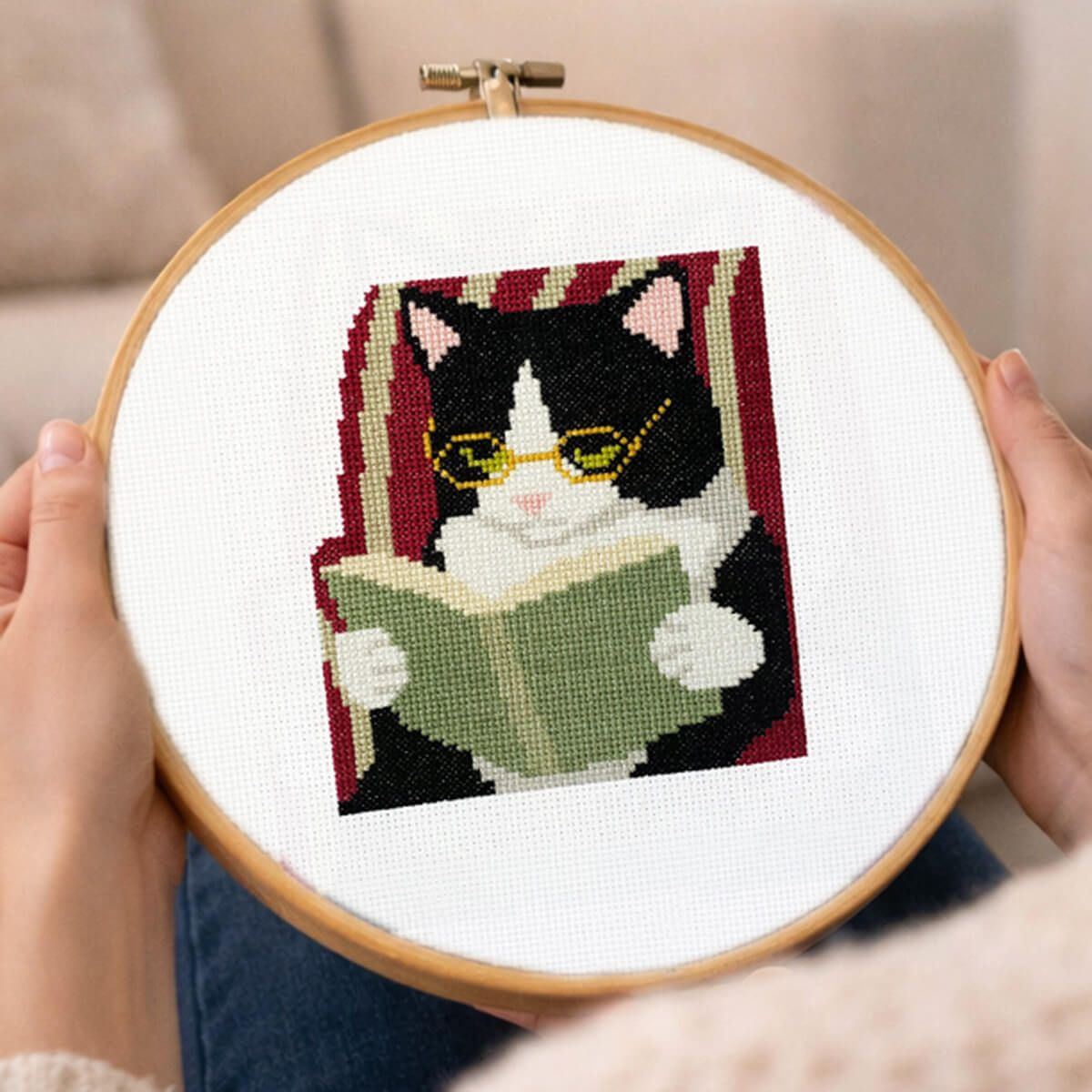 Cross-stitched design of a cat reading a book held in an embroidery hoop.
