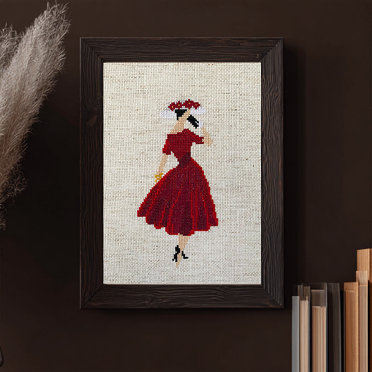 Framed cross-stitched artwork of a woman in a red dress on a dark wall.