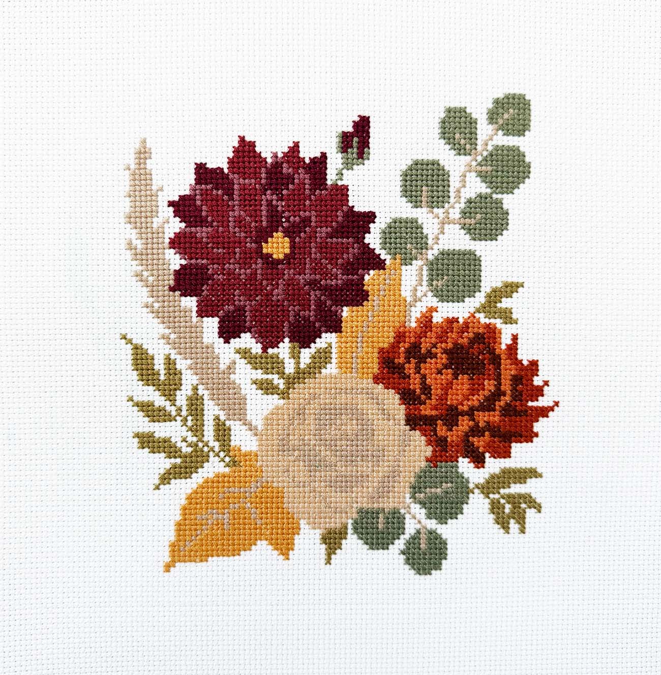 A cross-stitch floral design on white Aida fabric, featuring a burgundy dahlia, a red-orange marigold, a beige rose, and green foliage.