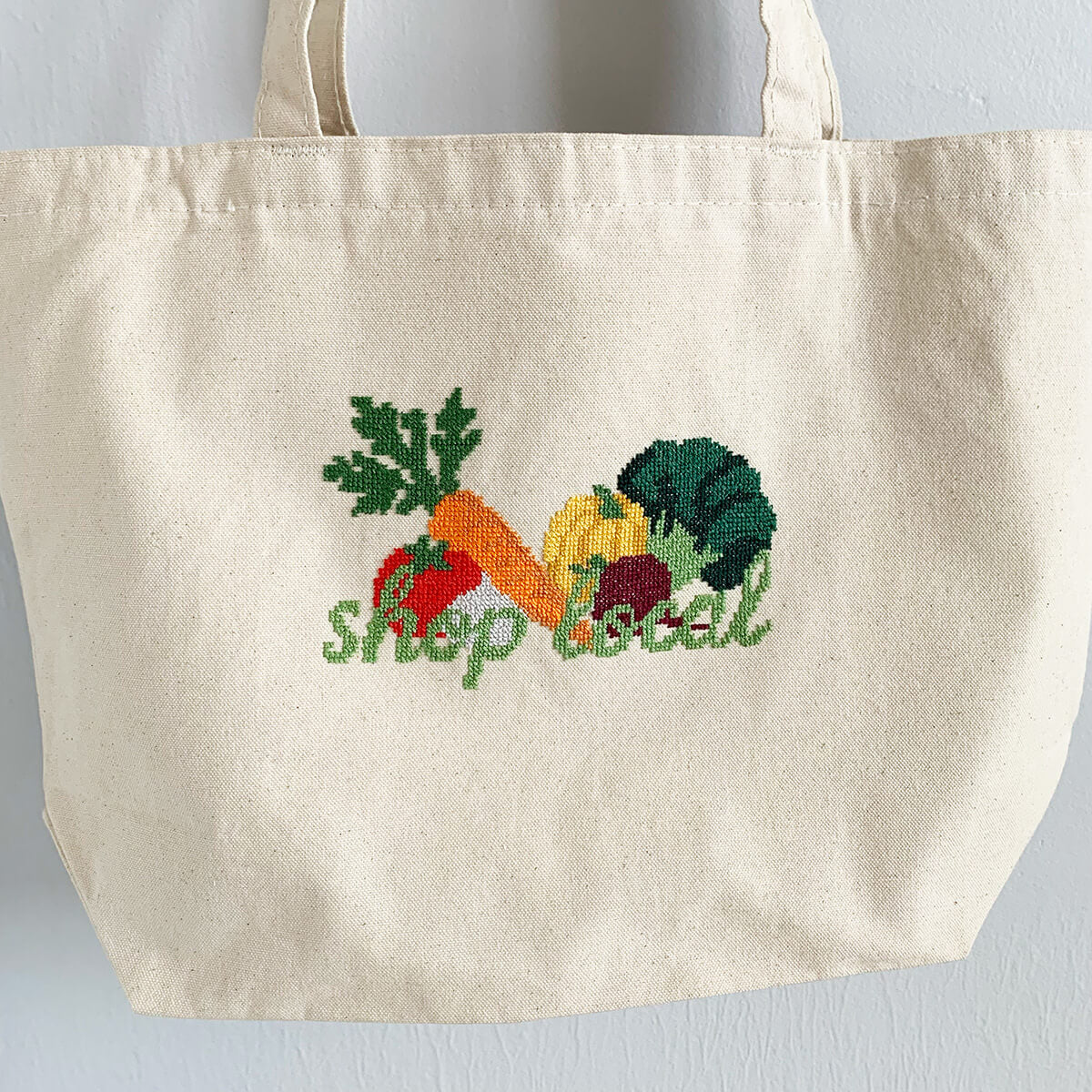 Beige tote bag with embroidered vegetables and 'shop local' text on a light gray background