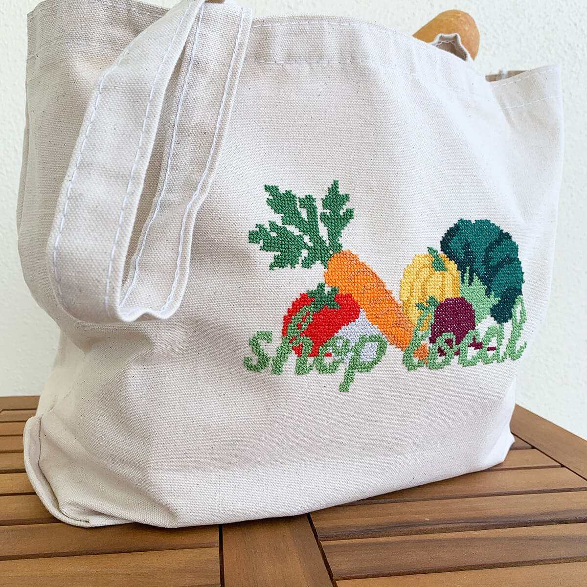Beige tote bag with embroidered vegetables and 'shop local' text on a wooden surface.