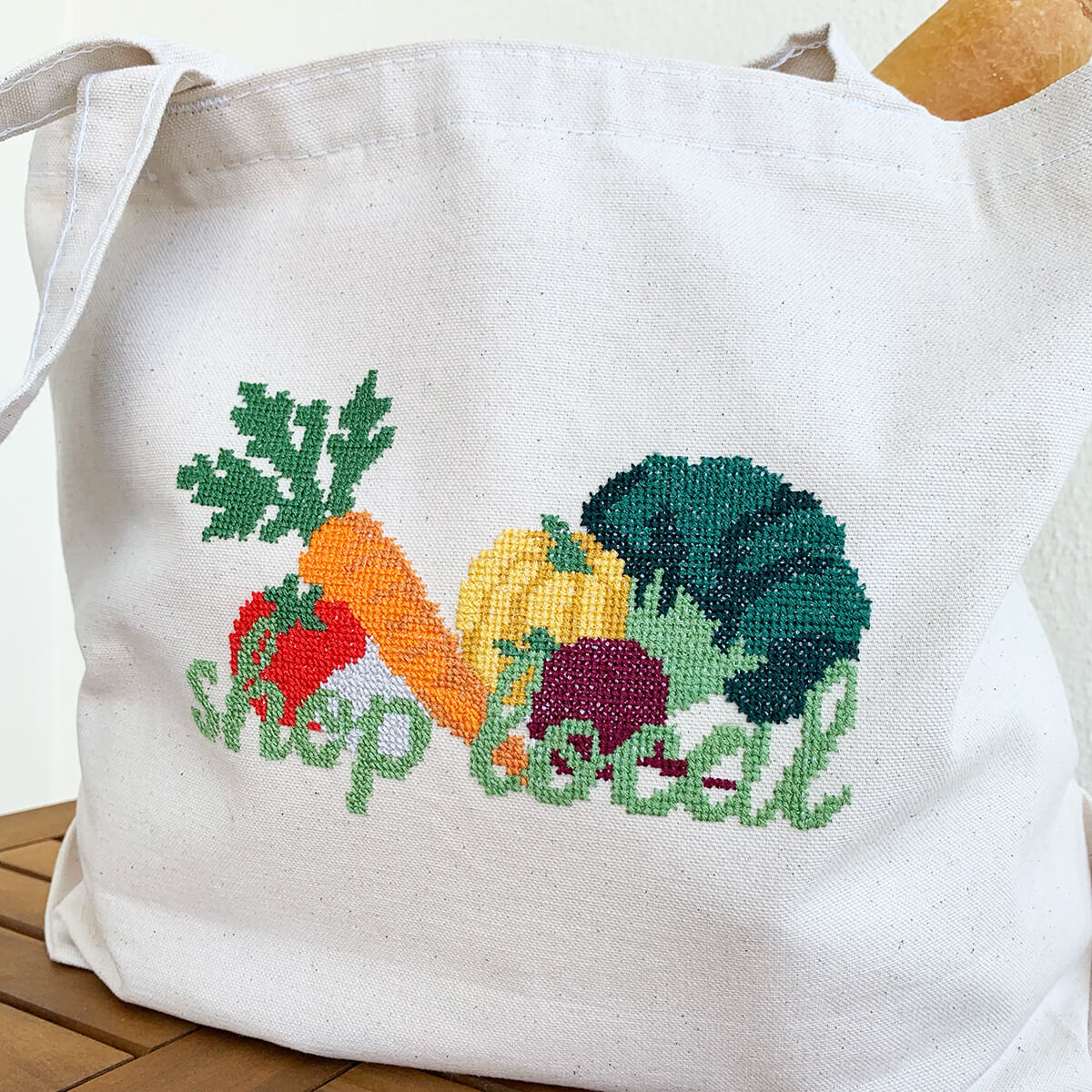 White tote bag with cross-stitched vegetables and 'shop local' text on a wooden surface.