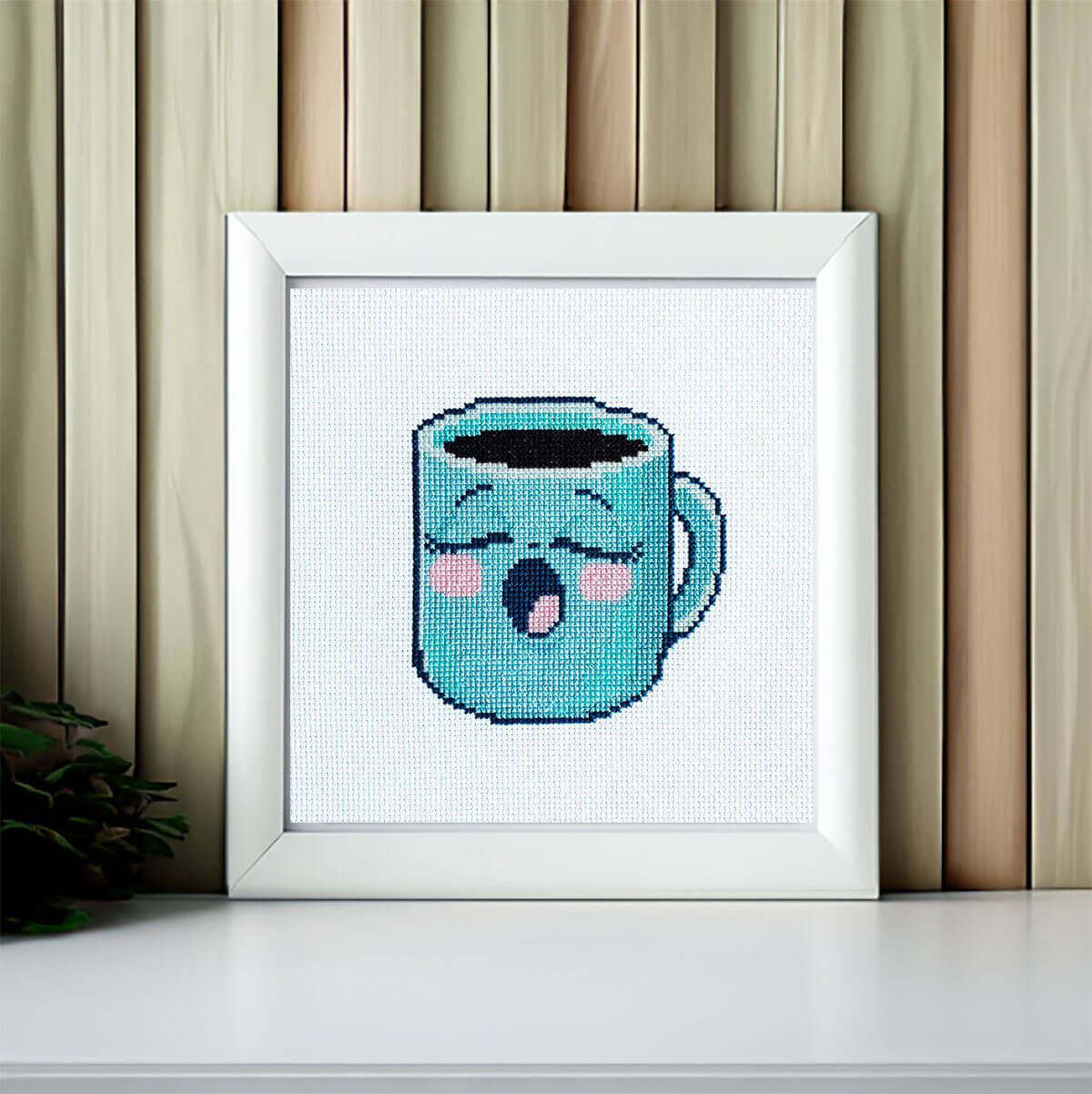 Framed cross-stitched artwork of a blue cup with a sleepy face on a white background.