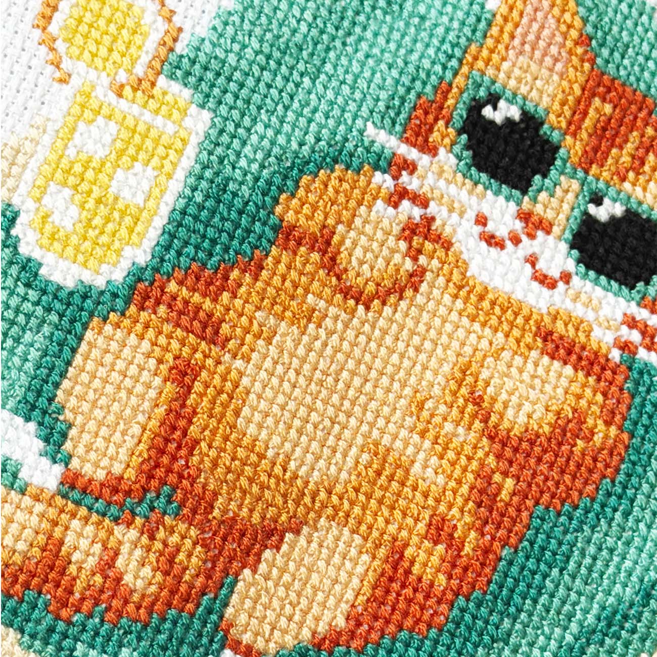 Close-up of a cross-stitch pattern depicting a cartoon cat wearing sunglasses, with a yellow cup on a teal background.
