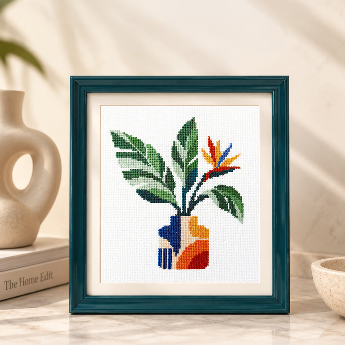 A framed cross-stitch artwork featuring a stylized green plant with a blue, orange, and red geometric vase, displayed on a marble surface with decorative props in a warm indoor setting.