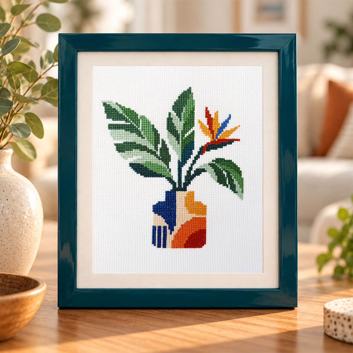 A framed cross-stitch artwork featuring a stylized plant with green leaves and a blue, orange, and yellow flower in a vase, displayed on a wooden table in a softly lit living room setting.