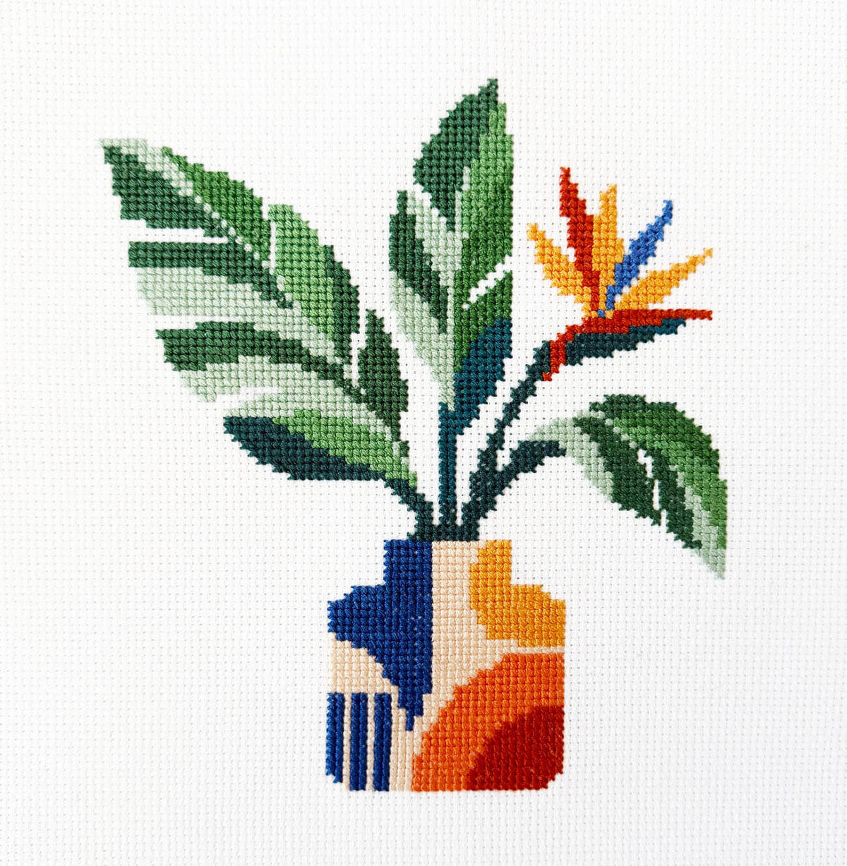 A full finished cross stitch design featuring a potted plant with green leaves and a bird of paradise flower, set against a white grid background.