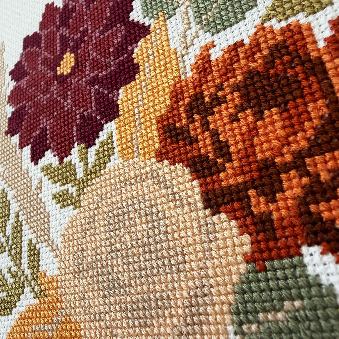 Close-up of a cross-stitch floral design showing textured stitched petals in burgundy, orange-brown, cream, yellow, and olive green on a white fabric ground.