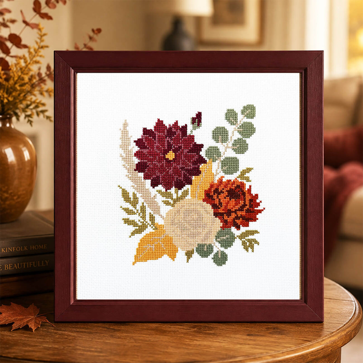A framed cross-stitch floral design displayed on a wooden table in a cozy living room setting. The artwork features a burgundy frame with a white fabric background and multicolored flowers including burgundy, orange, yellow, and green.