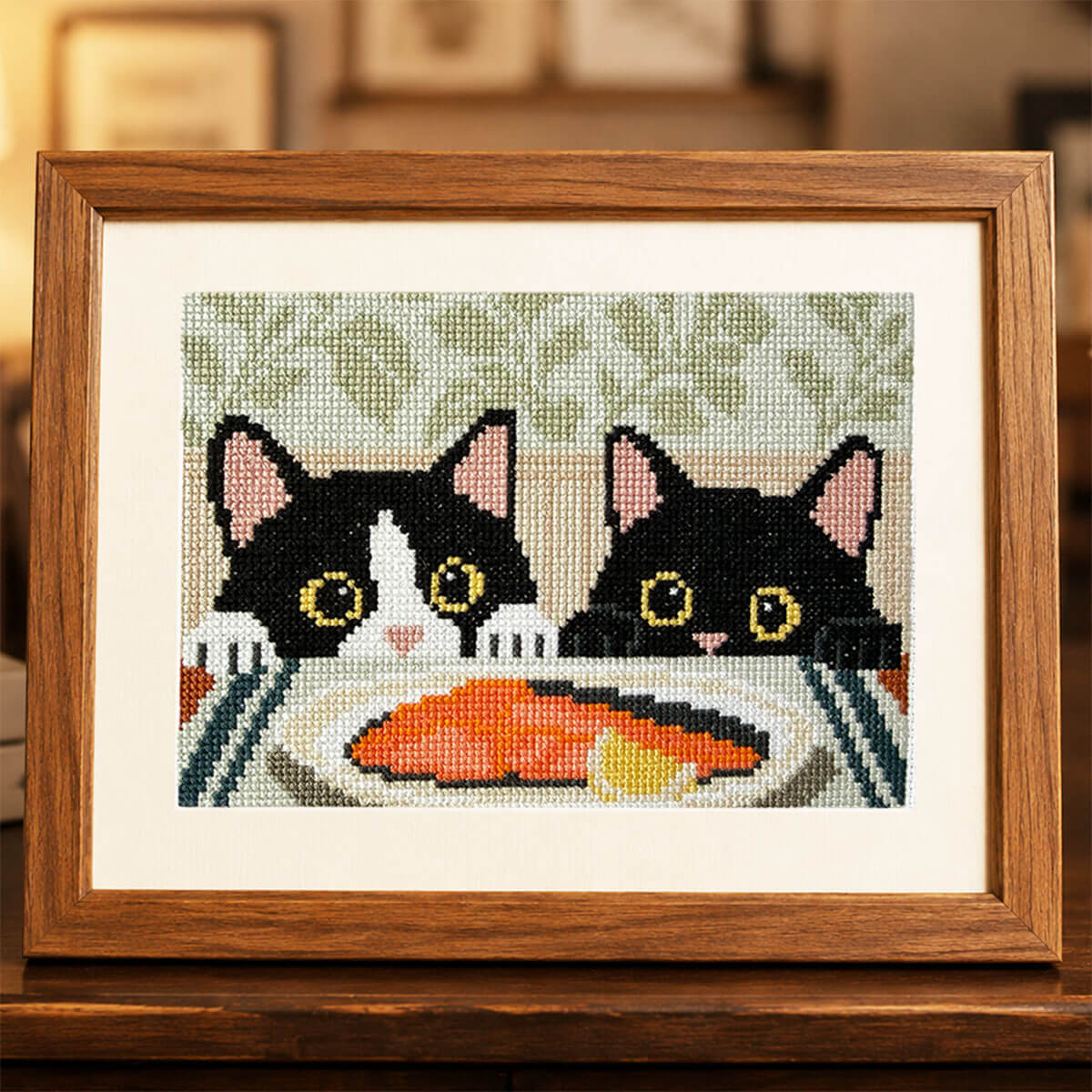 Framed Dinner Inspectors tuxedo cat counted cross stitch displayed on a dark wooden dining room console. This modern cat cross stitch kit features two curious black cats watching a salmon dinner in a warm, cozy home interior.