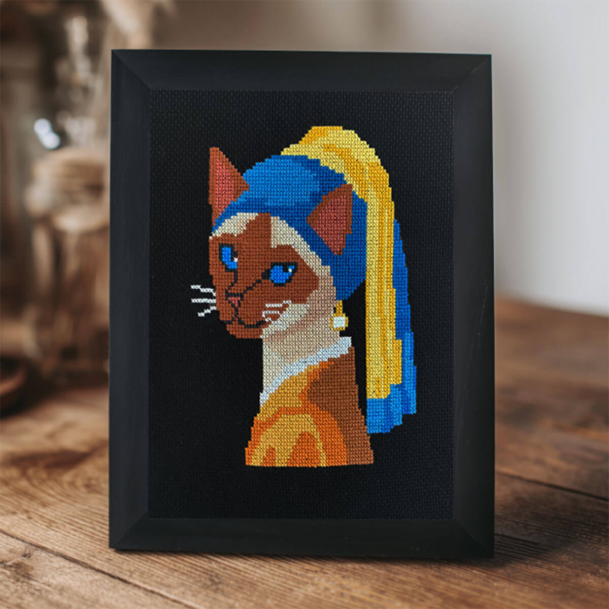 Framed cross-stitched art of a cat with a blue and yellow headband  on a wooden surface.