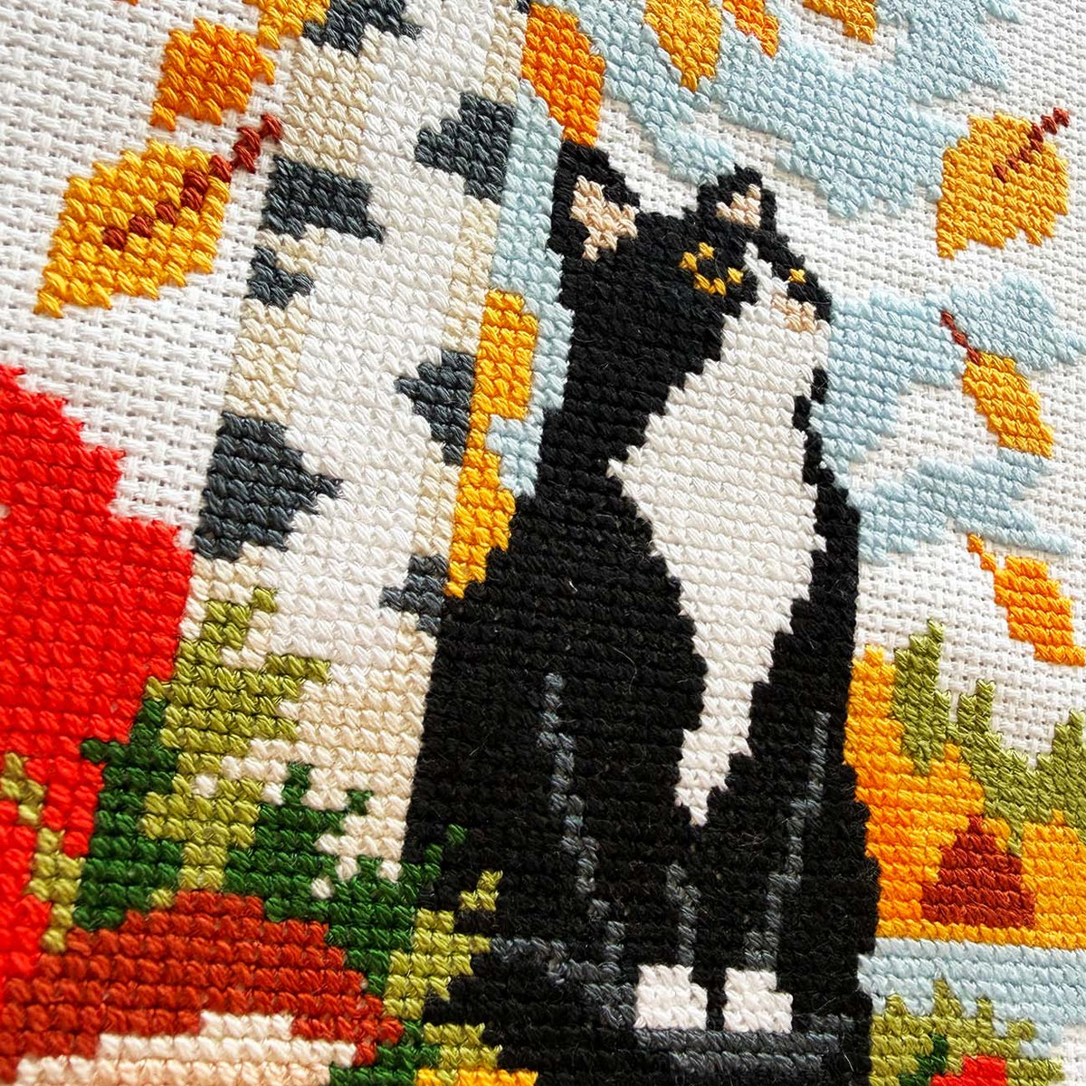 Close-up of a cross-stitch canvas featuring a black and white cat sitting in front of a birch tree with autumn leaves.