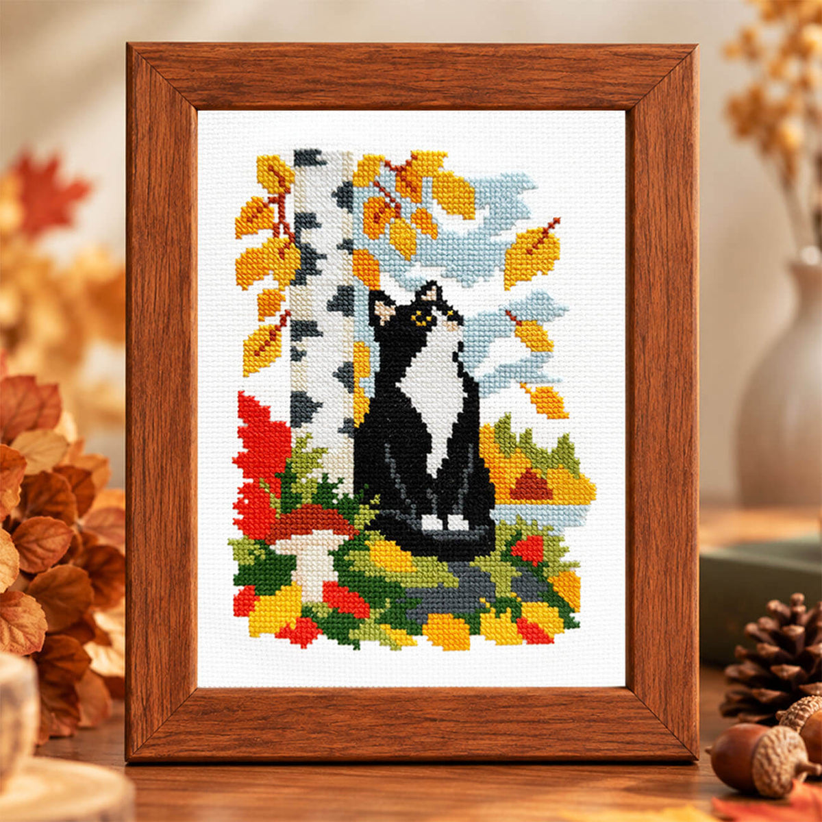 A framed cross-stitch artwork featuring a black-and-white cat sitting among autumn leaves and a birch tree, displayed in a warm wooden frame on a tabletop with seasonal decor around it.
