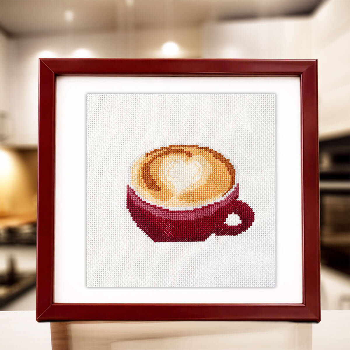 Framed cross-stitched picture of a coffee cup with a heart design on a blurred kitchen background