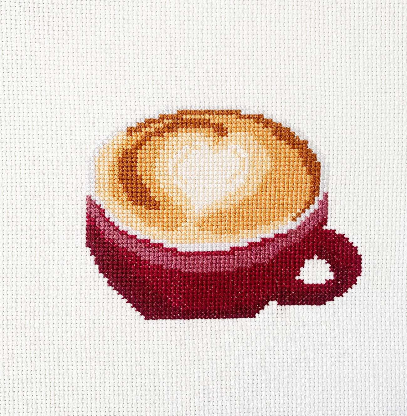 Latte with a heart foam design finished cross stitch piece on off-white Aida, showing full stitched design.