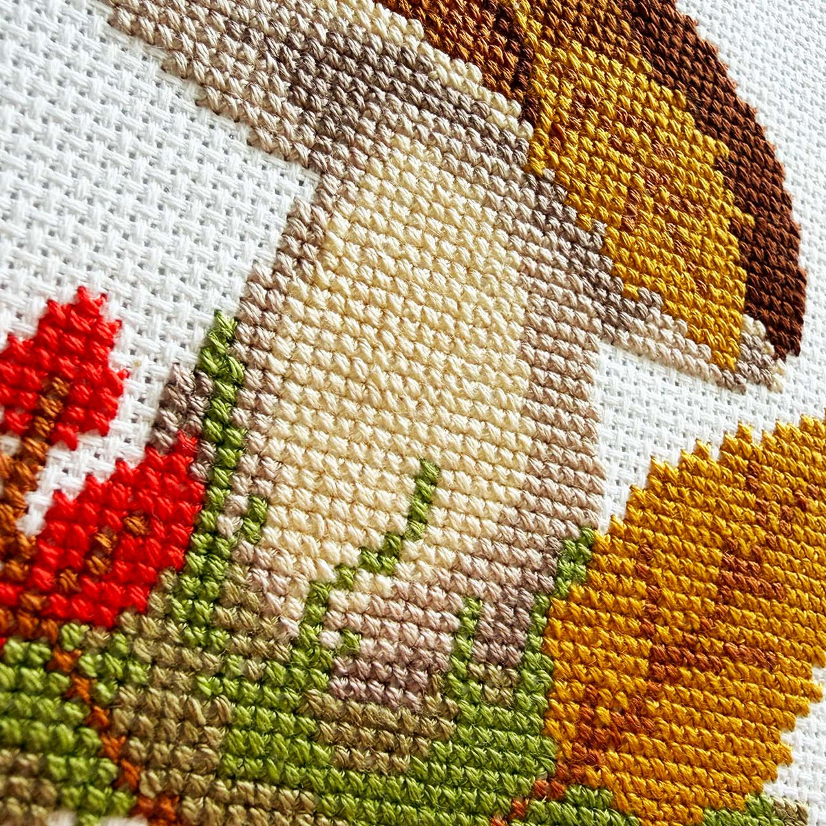 Close-up of a cross-stitch embroidery showing a mushroom with red and yellow autumn leaves, stitched on white Aida fabric.