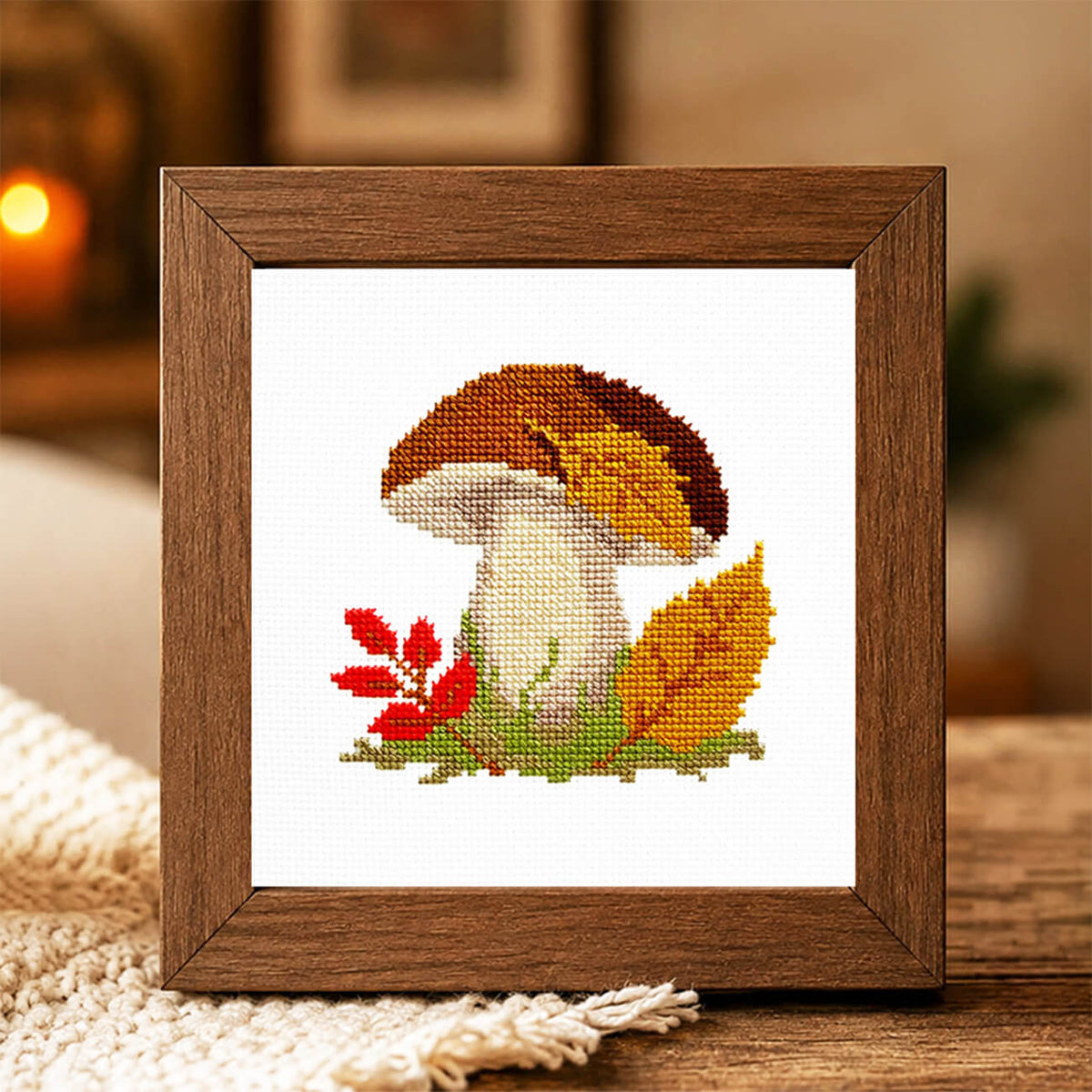 A framed cross-stitch artwork featuring a mushroom with autumn leaves, displayed on a wooden surface in a cozy indoor setting.