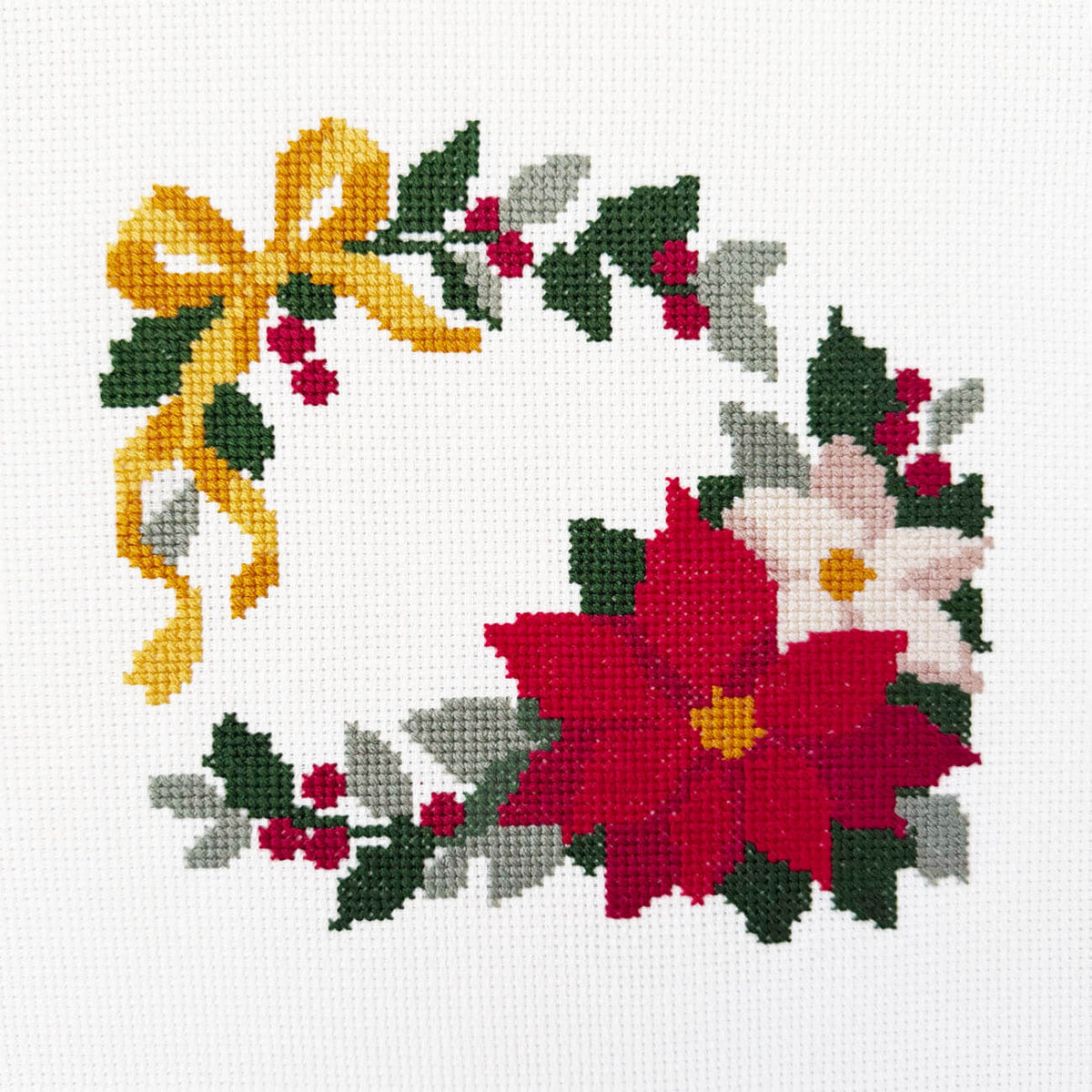 A festive cross-stitch wreath design featuring a large red poinsettia, a white flower, green leaves, red berries, and a yellow bow on a white fabric background.
