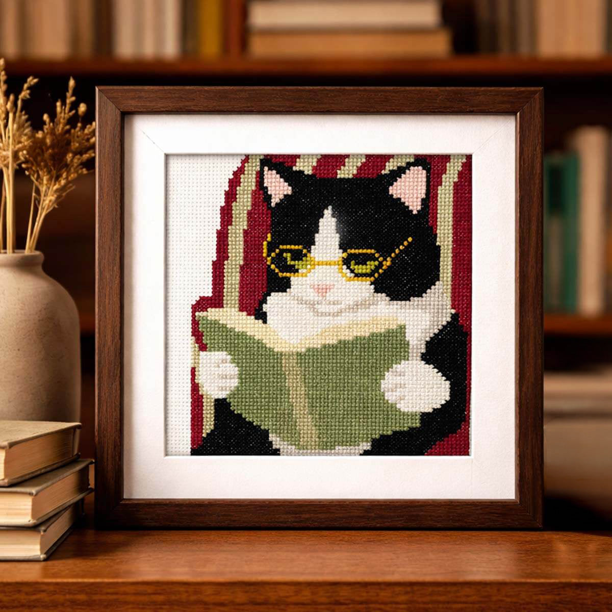 A framed cross-stitch artwork of a black-and-white cat reading a green book, displayed on a wooden shelf in a cozy room setting with books and a vase in the background.
