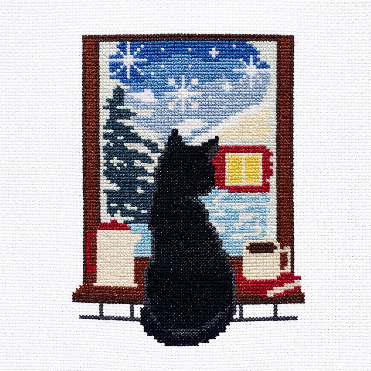 Cross-stitched picture of a black cat in front of a snowy cabin scene.