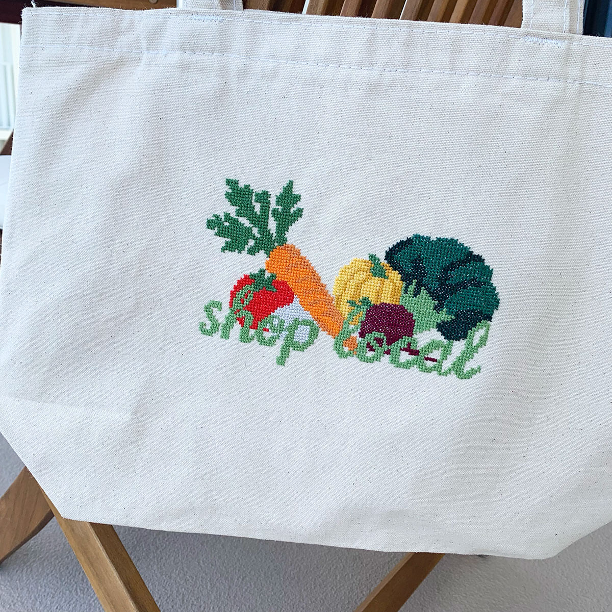 Beige tote bag with cross stitched vegetables and 'shop local' text on a wooden chair.